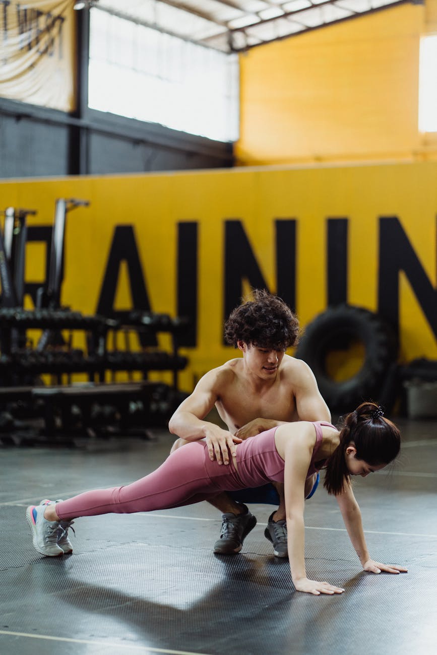 A personal trainer helps a woman with her plank exercise in a gym setting.